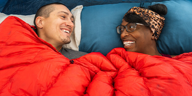 a mana and a women laying down in a red down enlightened equipment sleeping quilt