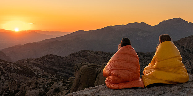 two women sitting on a mountain wrapped in a orange an yellow enlightened equipment sleeping quilt