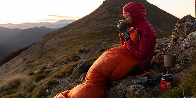 a picture of a women drinking coffee while being wrapped in a salmon orange sleeping quilt