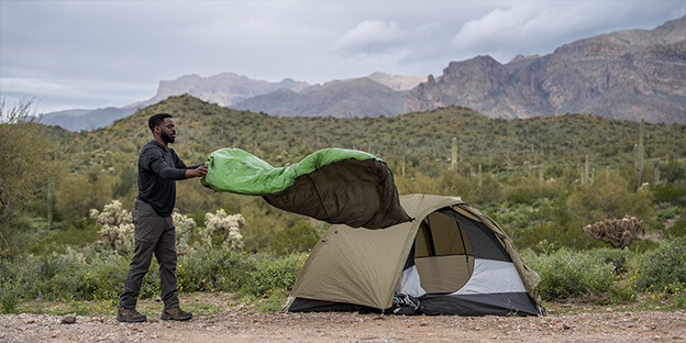 a man holding a forest color synthetic quilt infront of a tent in the desert